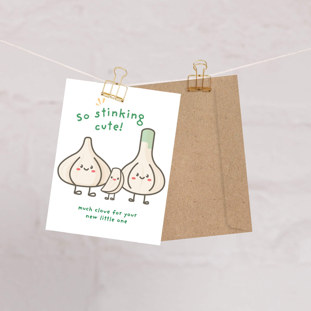 A funny baby shower card with a cute garlic illustration and the text 'so stinking cute!' hangs from a string with a gold binder clip, with a brown kraft envelope behind it.