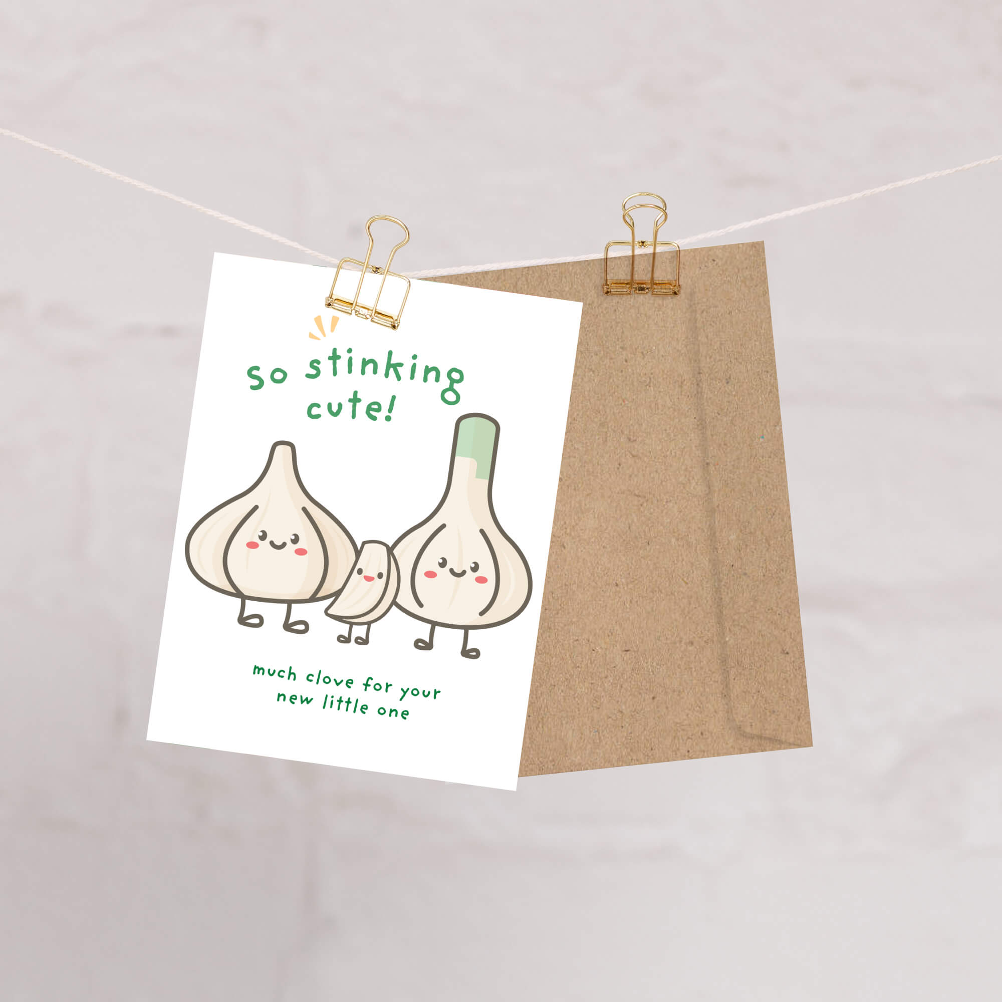 A funny baby shower card with a cute garlic illustration and the text 'so stinking cute!' hangs from a string with a gold binder clip, with a brown kraft envelope behind it.