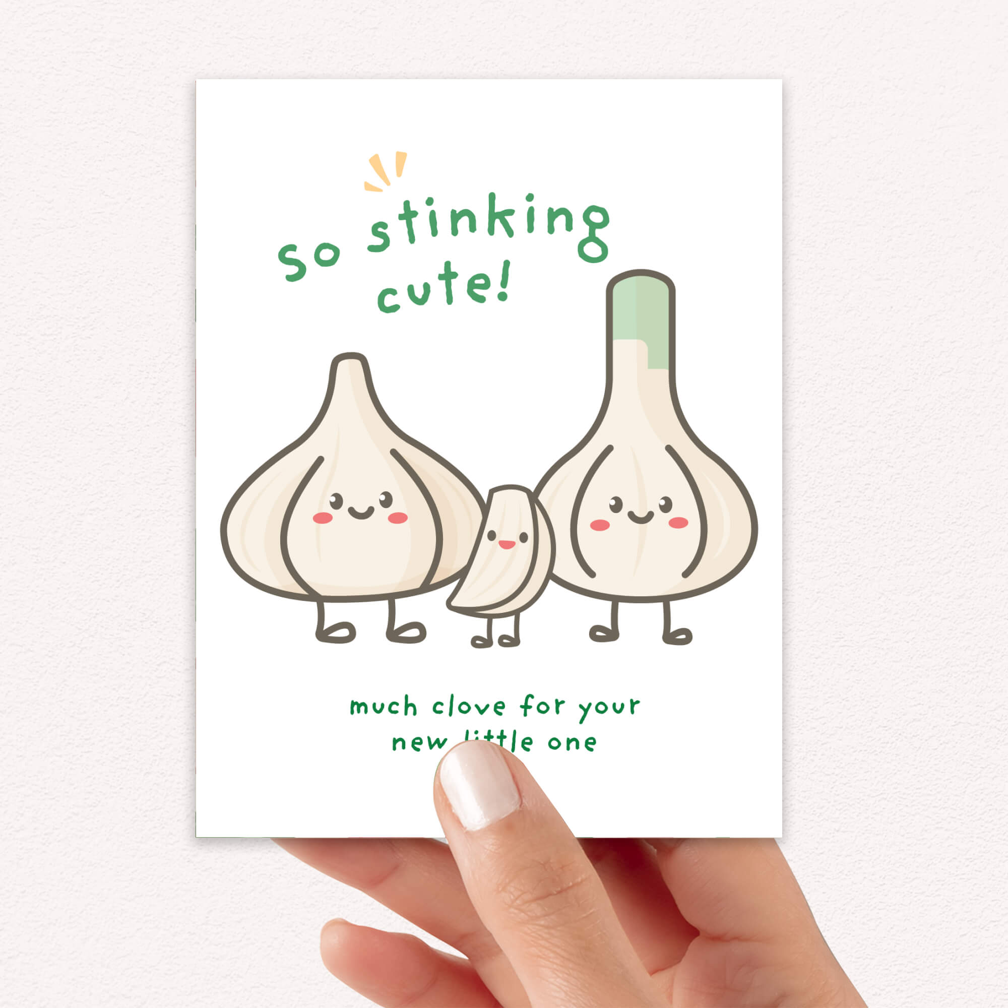 A hand holds a new baby card featuring a cute illustration of three smiling garlic cloves with the text 'so stinking cute!' and 'much clove for your new little one' in green lettering.
