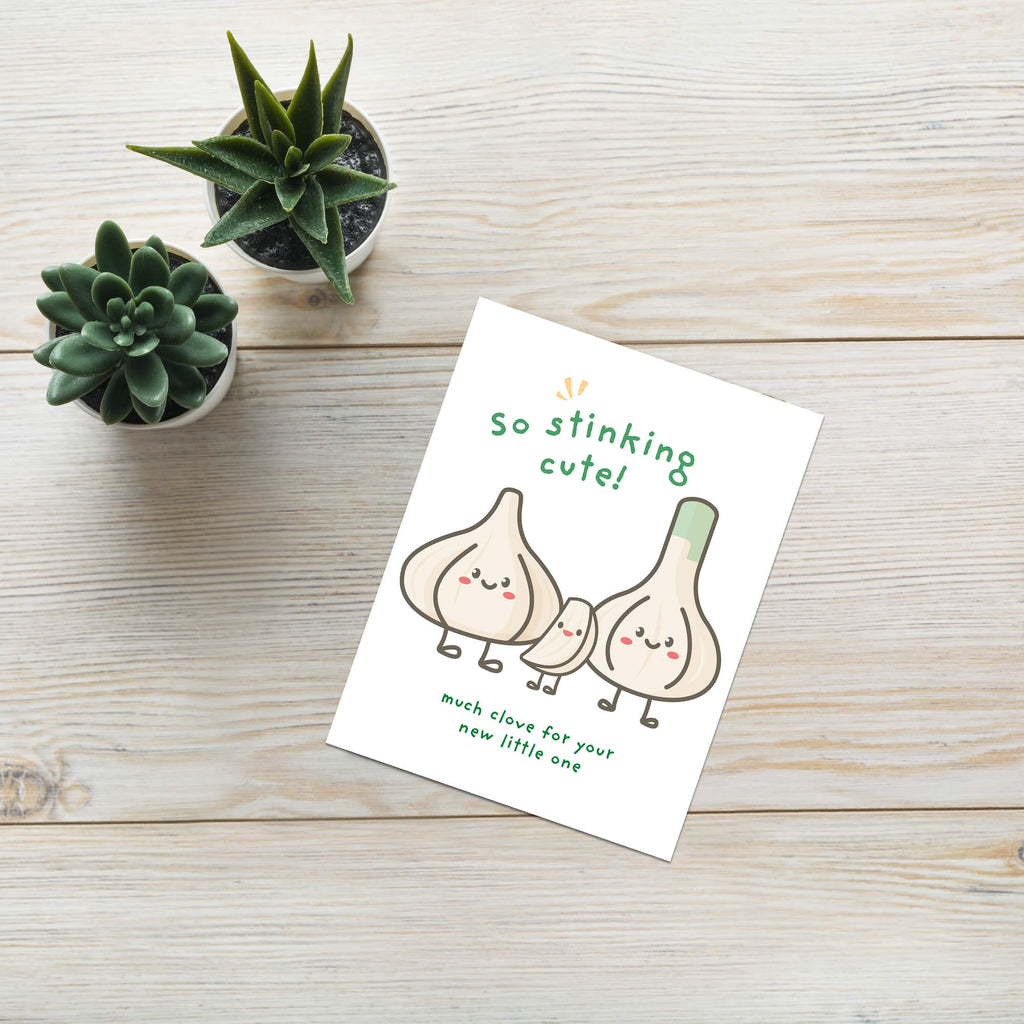 A funny baby shower card featuring a cute cartoon illustration of three garlic bulbs with faces and the text 'so stinking cute!' and 'much love for your new little one', placed on a light wood surface next to two small potted succulents.
