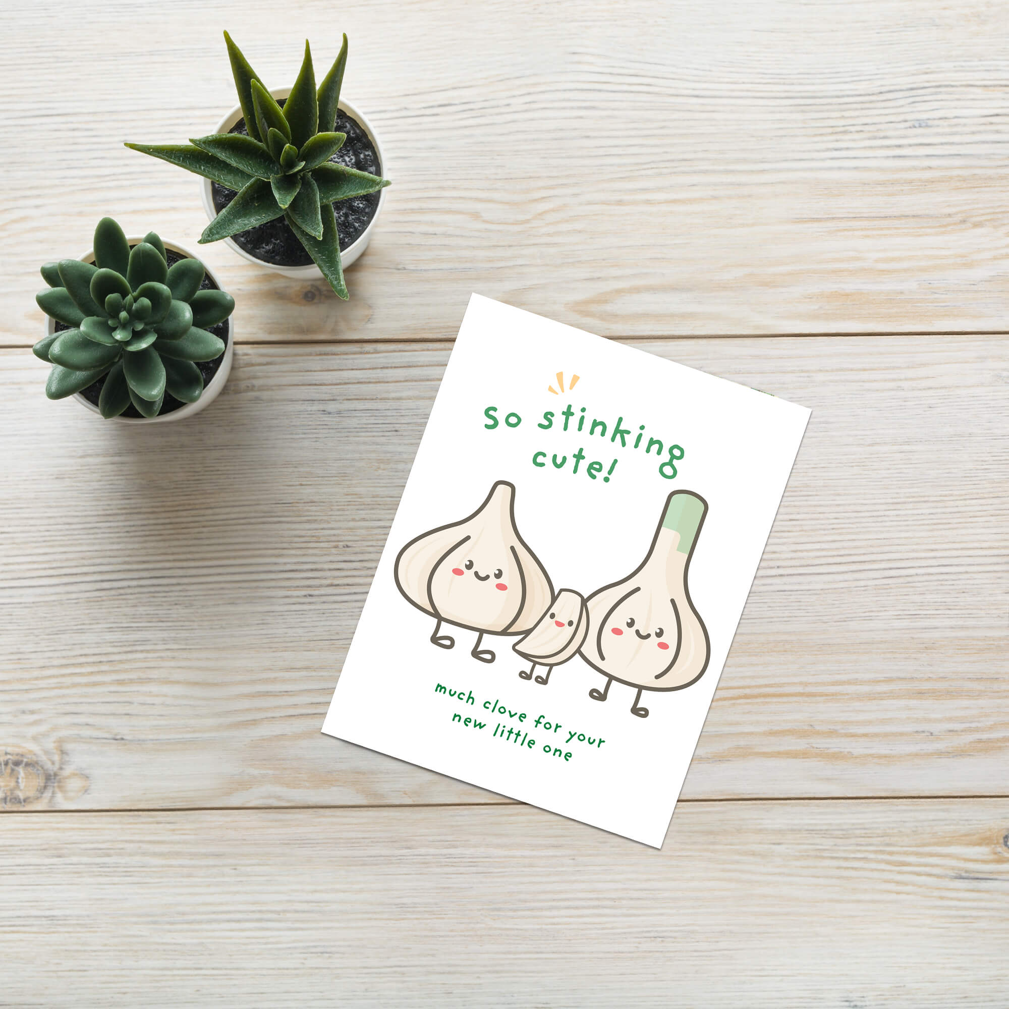 A funny baby shower card featuring a cute cartoon illustration of three garlic bulbs with faces and the text 'so stinking cute!' and 'much love for your new little one', placed on a light wood surface next to two small potted succulents.