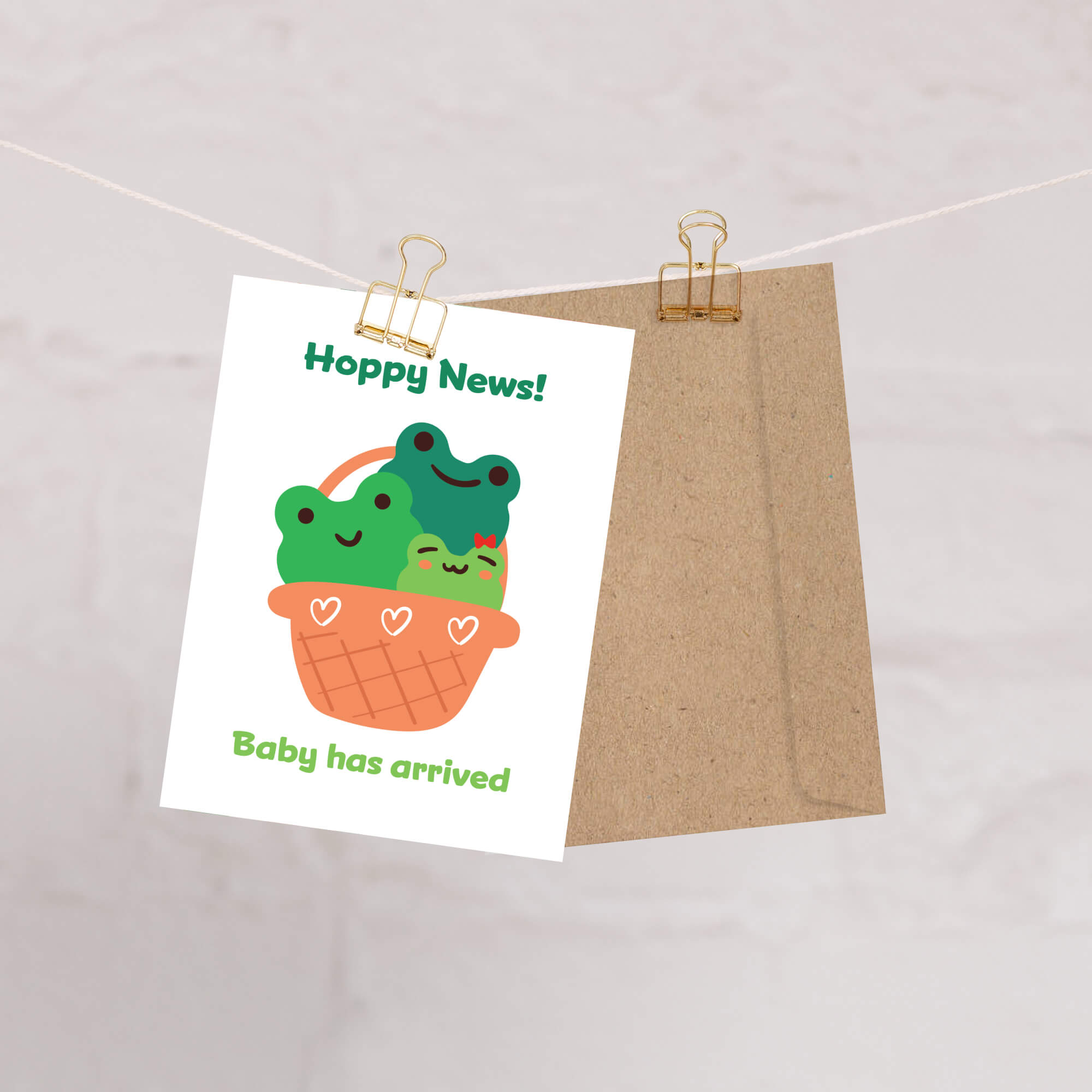 A green new baby card clipped to a string with a kraft envelope behind it, featuring a cute cartoon frog and baby bird in a basket with the text 'Hoppy News!' and 'Baby has arrived'.