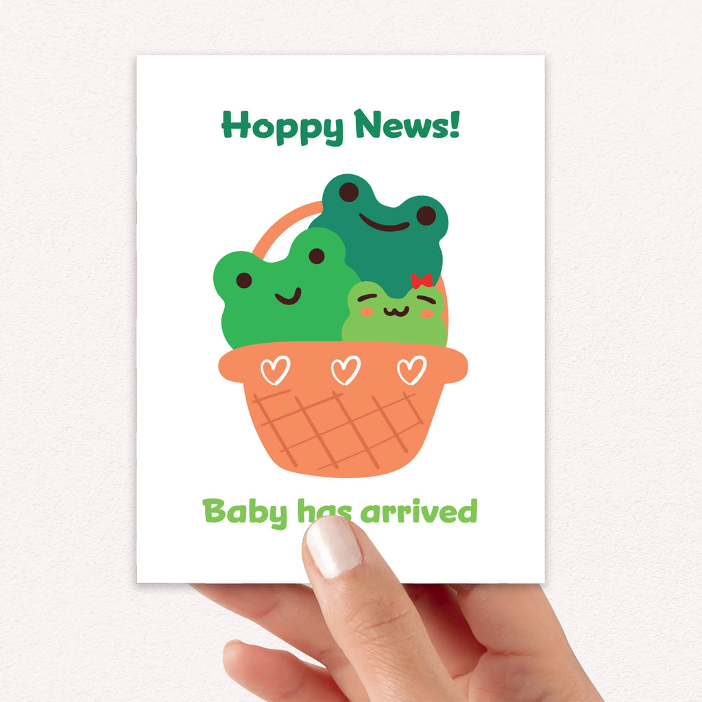 A hand holding a new baby shower card that reads 'Hoppy News!' and 'Baby has arrived' with a cute illustration of three smiling green frogs in an orange basket.