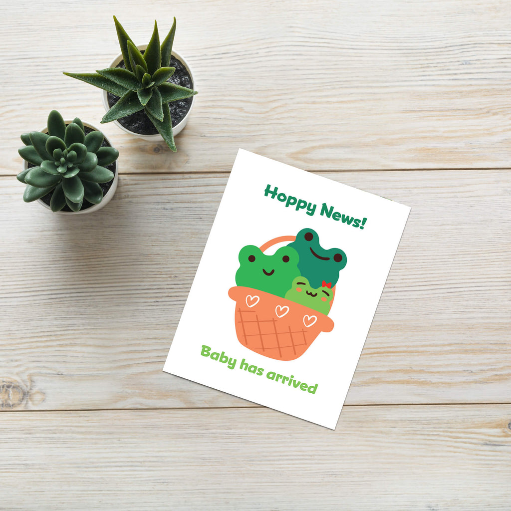 A frog inspired baby shower card with a cute illustration of two green frog babies in an orange basket, placed on a light wood tabletop next to two small potted succulents. The card reads 'Hoppy News!' and 'Baby has arrived'.
