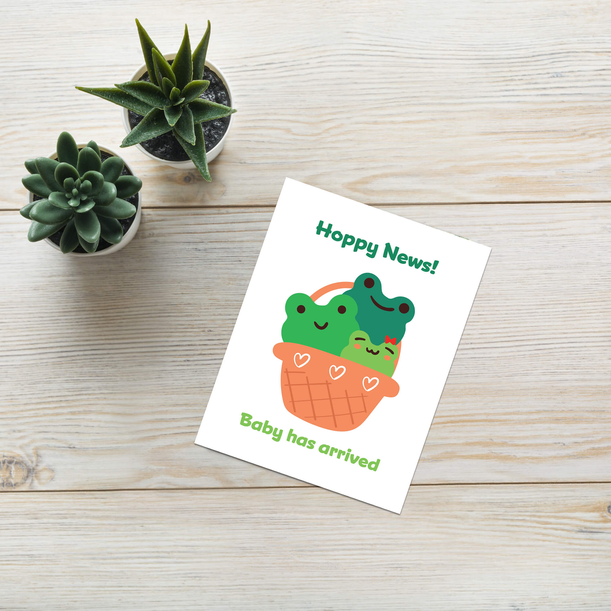 A frog inspired baby shower card with a cute illustration of two green frog babies in an orange basket, placed on a light wood tabletop next to two small potted succulents. The card reads 'Hoppy News!' and 'Baby has arrived'.