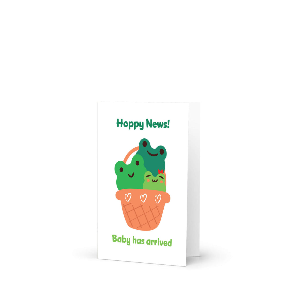 A baby shower card with a cute illustration of three green cartoon frogs in an orange basket, with the text 'Hoppy News!' at the top and 'Baby has arrived' at the bottom, shown on a white background.