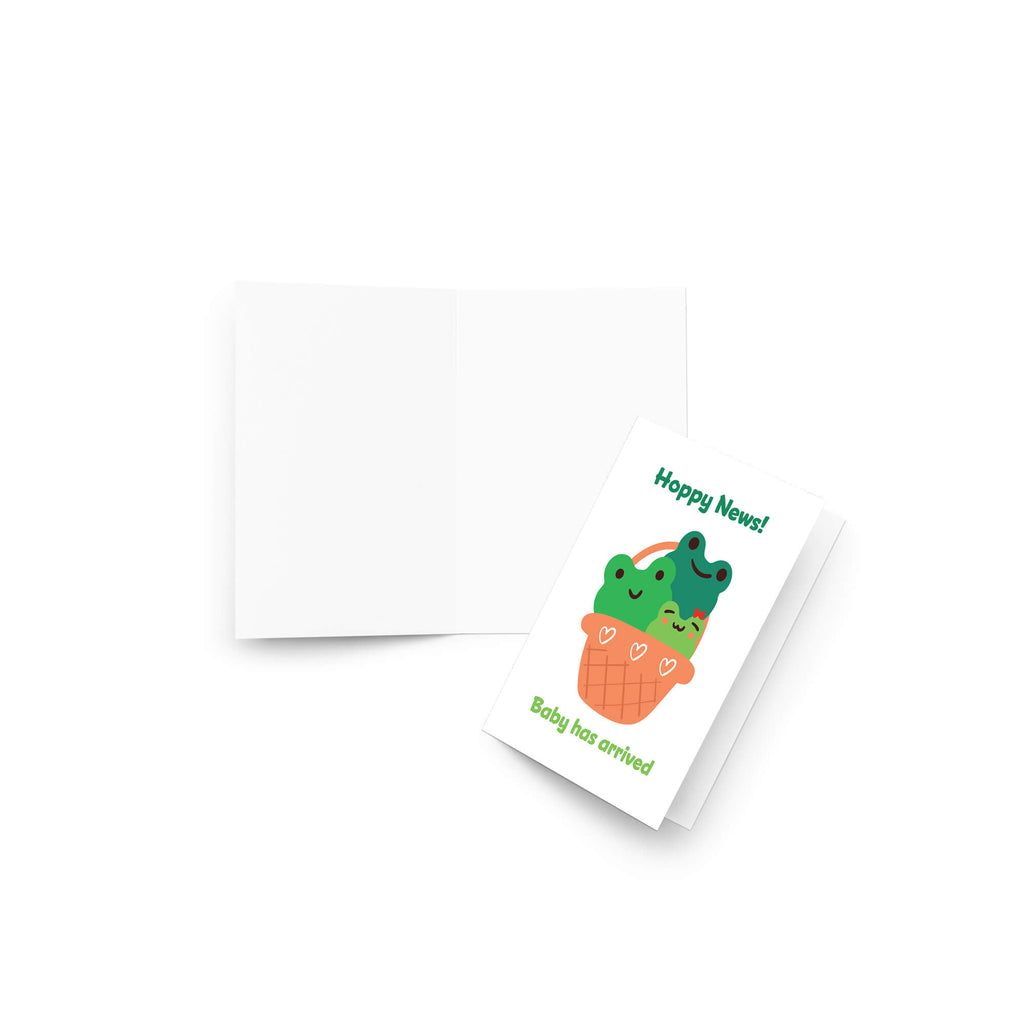 A folded new baby shower card with a cute cartoon illustration of two green frogs in an orange basket, shown next to its matching white envelope on a clean white background.