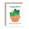 A baby shower card with a brown kraft envelope behind it, featuring a cute illustration of three smiling frogs in an orange basket with the text 'Hoppy News!' at the top and 'Baby has arrived' at the bottom.