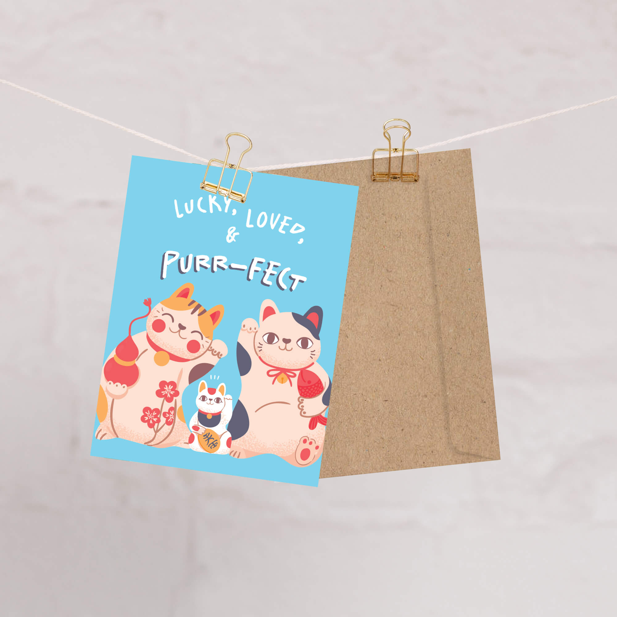 A Japanese new baby greeting card with a light blue background and cartoon cats, clipped to a string with a kraft paper envelope behind it on a light marble-like surface.