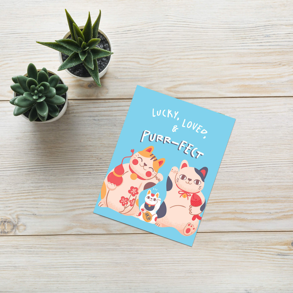 A cat baby shower card featuring three cute illustrated cats and the text 'LUCKY, LOVED & PURR-FELT' lies on a light wood surface next to two small potted succulents.