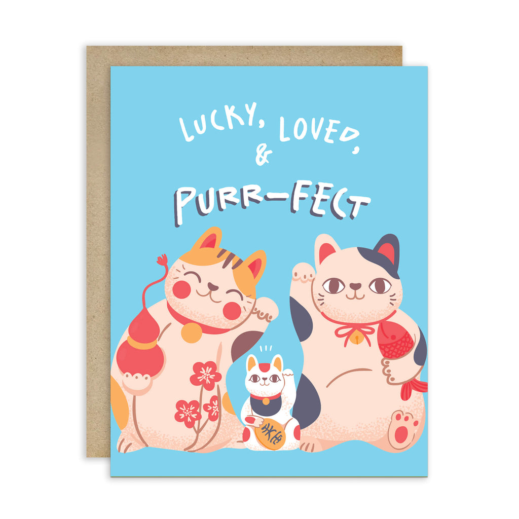 A Japanese baby shower greeting card with a light blue background and a kraft envelope behind it. The card features the text 'LUCKY, LOVED, & PURR-FECT' above three cute illustrated maneki-neko cats in peach, orange, white, and black with red accents.