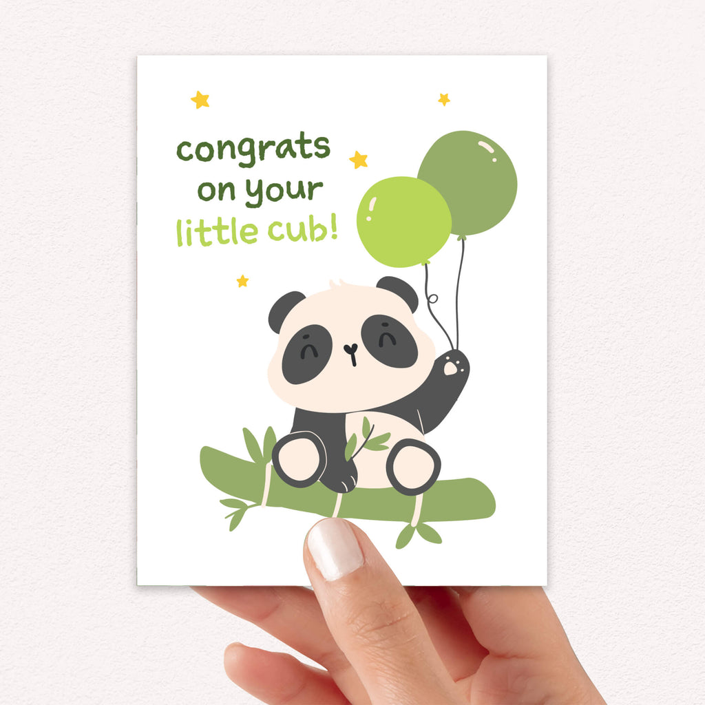A hand holding a baby congratulations card featuring a cute cartoon panda cub holding green balloons on a bamboo branch, with the text 'congrats on your little cub!'.
