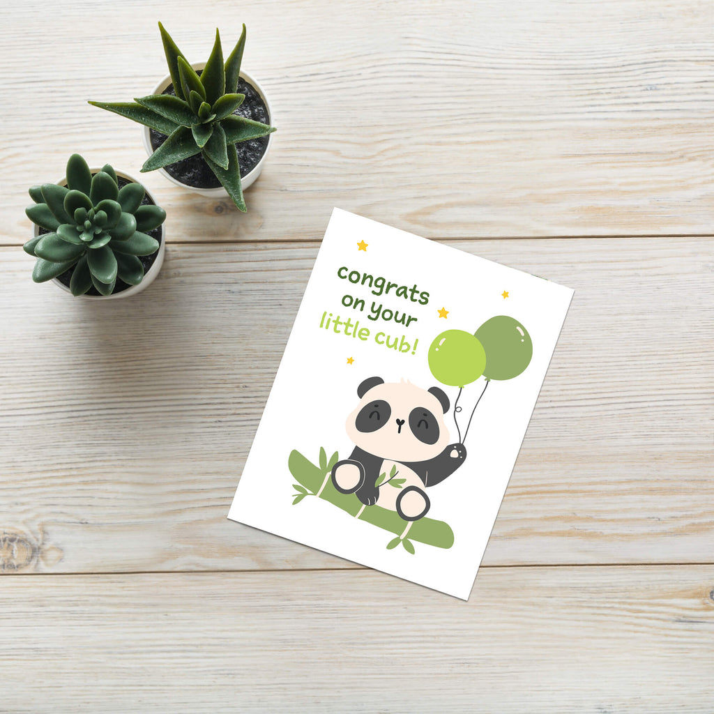 A new baby greeting card featuring a cute cartoon panda cub holding green balloons, with the text 'congrats on your little cub!'. The card is styled on a light wood background next to two small potted succulents.