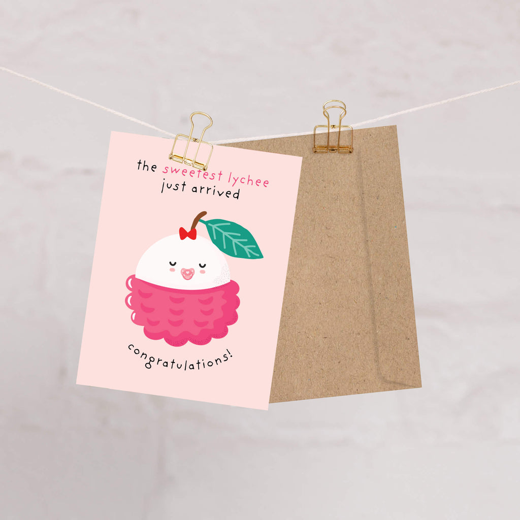 A pink new baby card clipped to a string with a kraft paper envelope behind it. The card features a cute cartoon lychee illustration with the text 'the sweetest lychee just arrived' and 'congratulations!'.