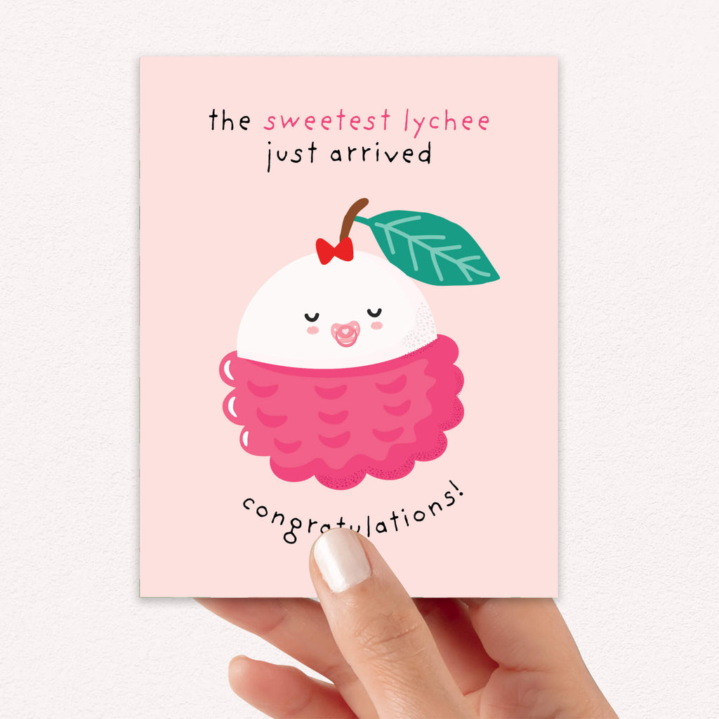 A hand holding a pink new baby card featuring a cute cartoon lychee illustration with the text 'the sweetest lychee just arrived' and 'congratulations!'.