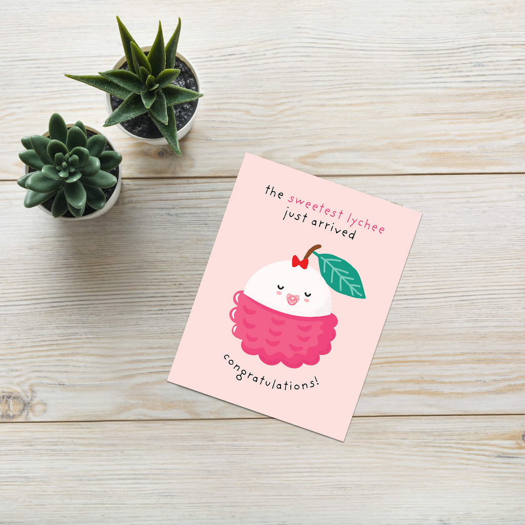 A pink congratulations baby shower card featuring a cute cartoon lychee illustration, styled on a light wood tabletop next to two small potted succulents.