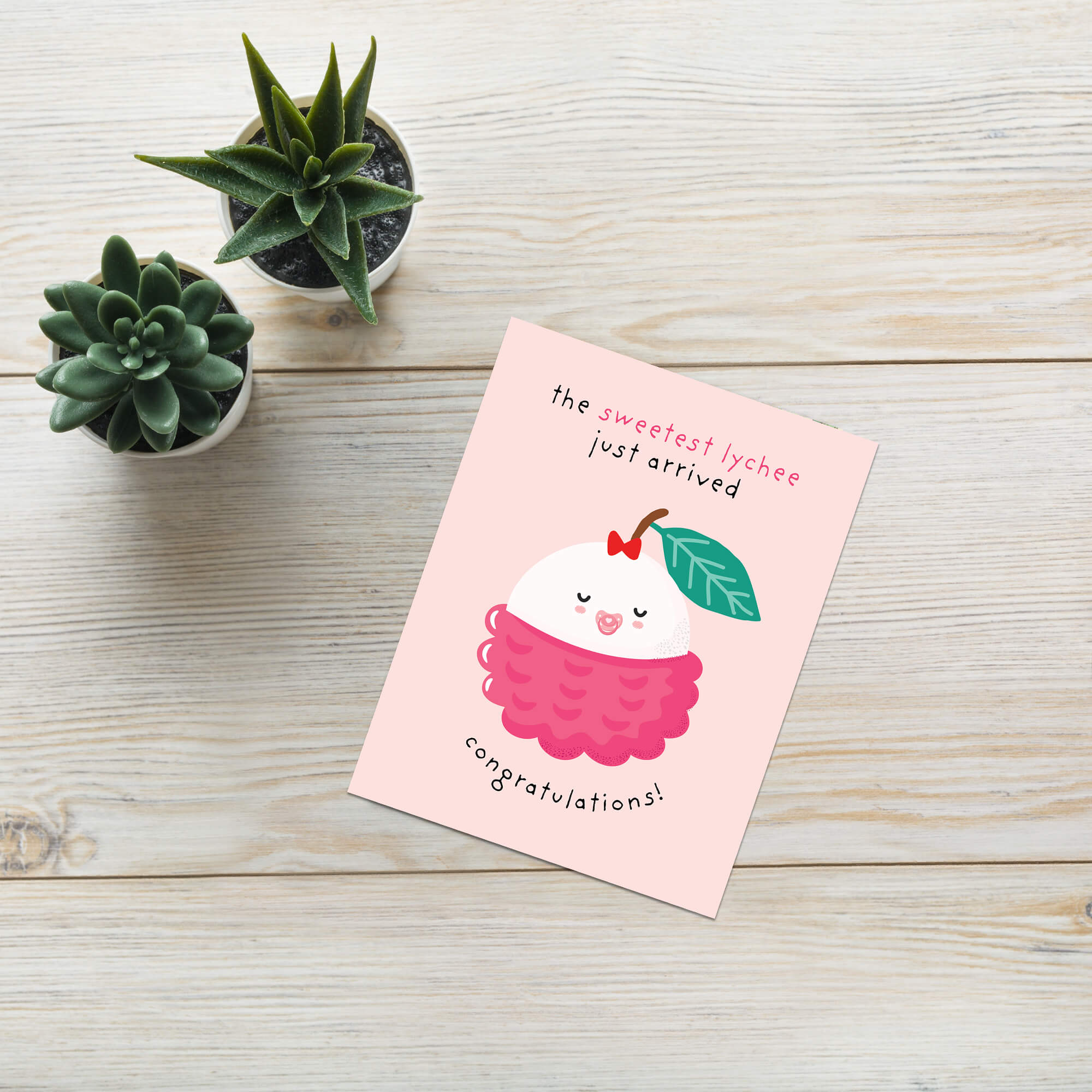 A pink congratulations baby shower card featuring a cute cartoon lychee illustration, styled on a light wood tabletop next to two small potted succulents.