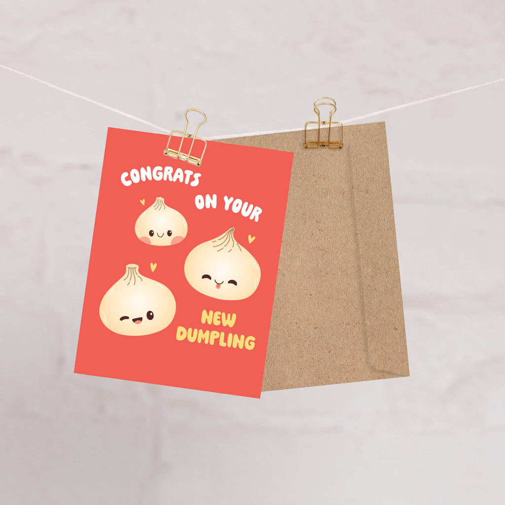 A red greeting card with cute dumpling illustrations and the text 'CONGRATS ON YOUR NEW DUMBLING' is clipped to a string next to a brown kraft envelope on a light marble background.