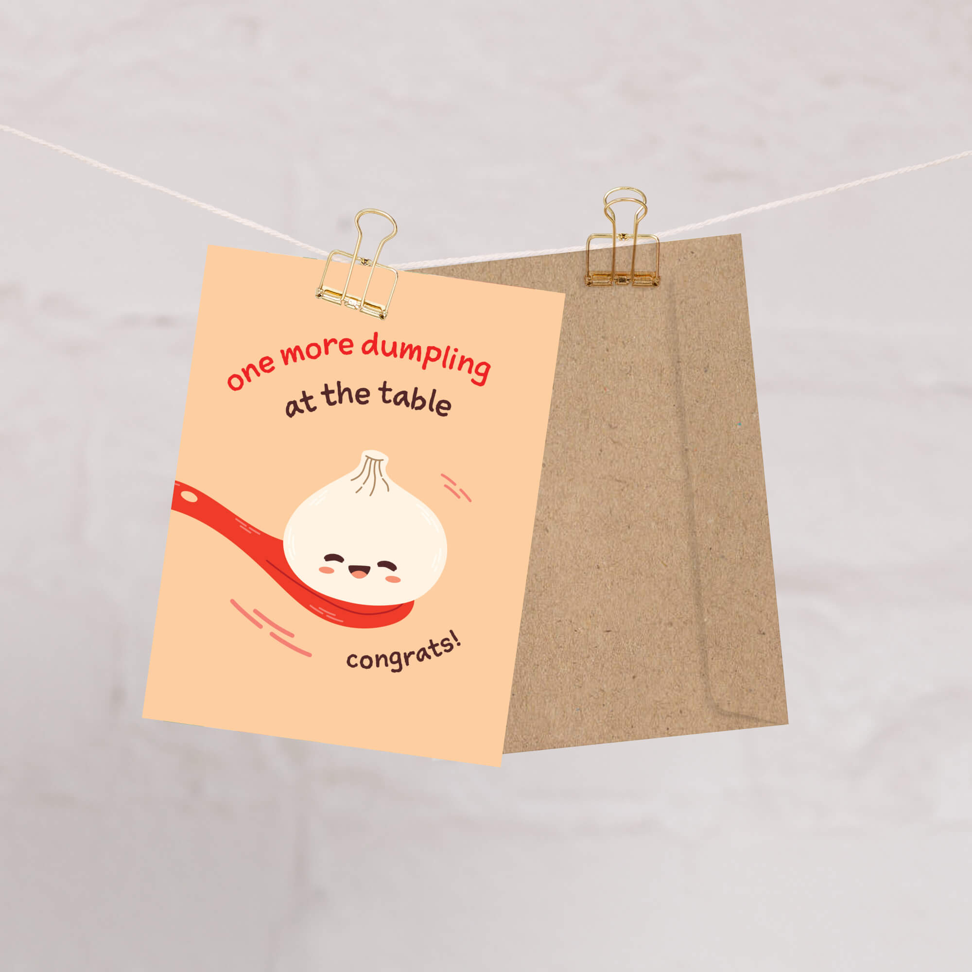 A cute dumpling-themed congratulations newborn card with a peach background, featuring a smiling dumpling on a red spoon and the text 'one more dumpling at the table' and 'congrats!'. The card is shown hanging from a wire with two binder clips, with a brown kraft envelope behind it on a light marble background.
