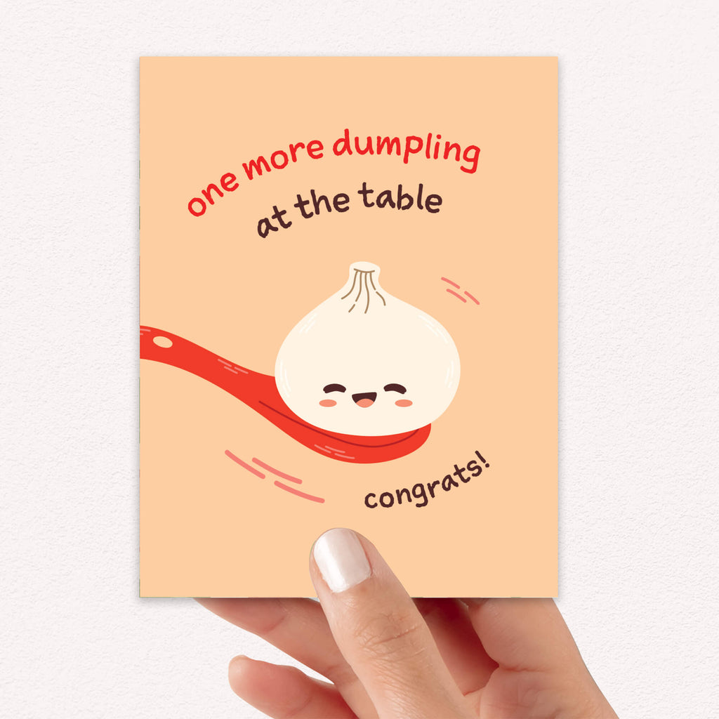 A hand holding a peach-colored greeting card with a cute cartoon dumpling on a red spoon and the text 'one more dumpling at the table' and 'congrats!'.
