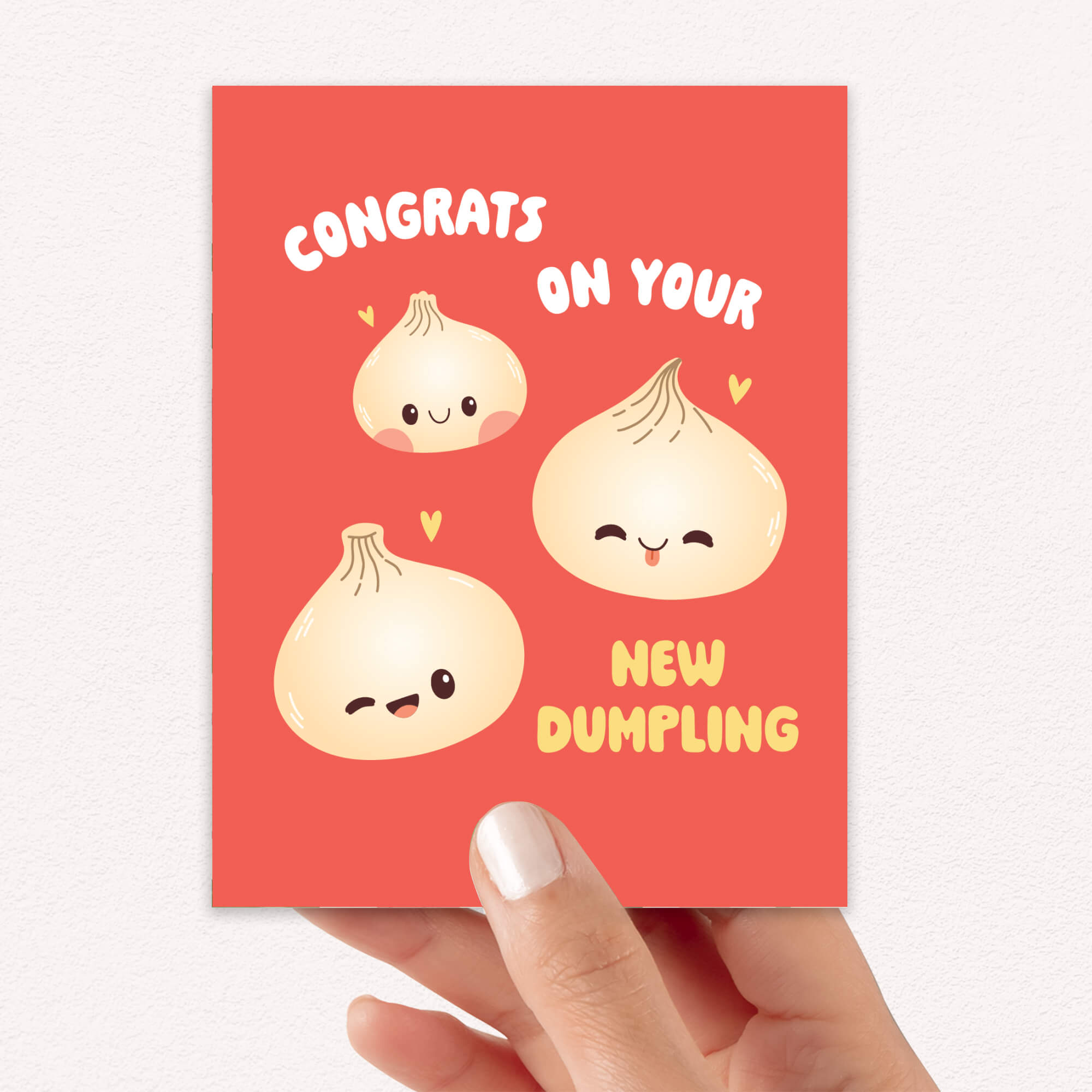 A hand holding a red greeting card with cute cartoon dumpling faces and the text 'CONGRATS ON YOUR NEW DUMBLING' on a light textured background.