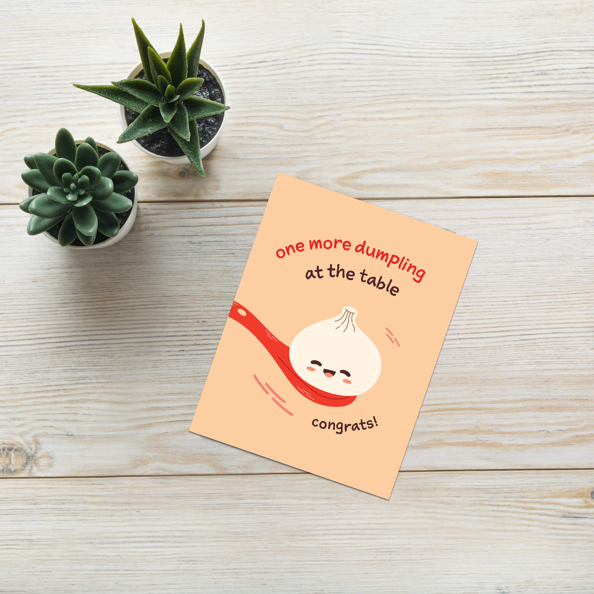 A new baby greeting card with a peach background, featuring a cartoon dumpling on a red spoon and the text 'one more dumpling at the table' and 'congrats!', placed on a light wood surface next to two small potted succulents.