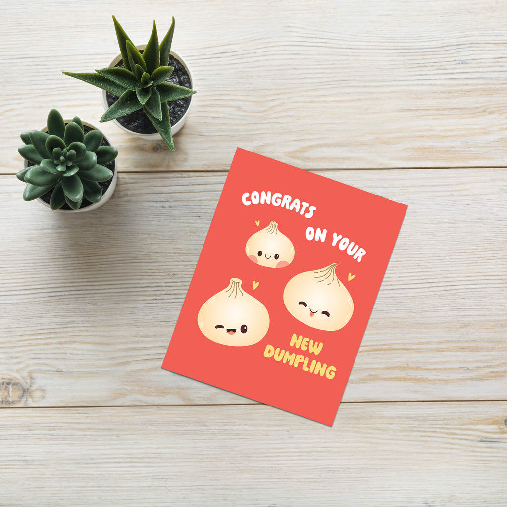 A red greeting card with cute cartoon dumplings and the text 'CONGRATS ON YOUR NEW DUMpLING' is displayed on a light wood surface next to two small potted succulents.