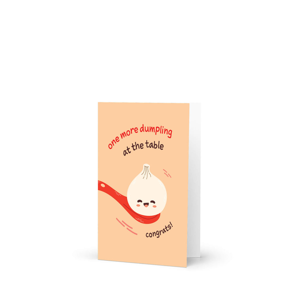 A cute baby shower card with a peach background featuring a cute cartoon dumpling on a red spoon and the text 'one more dumpling at the table' and 'congrats!'.