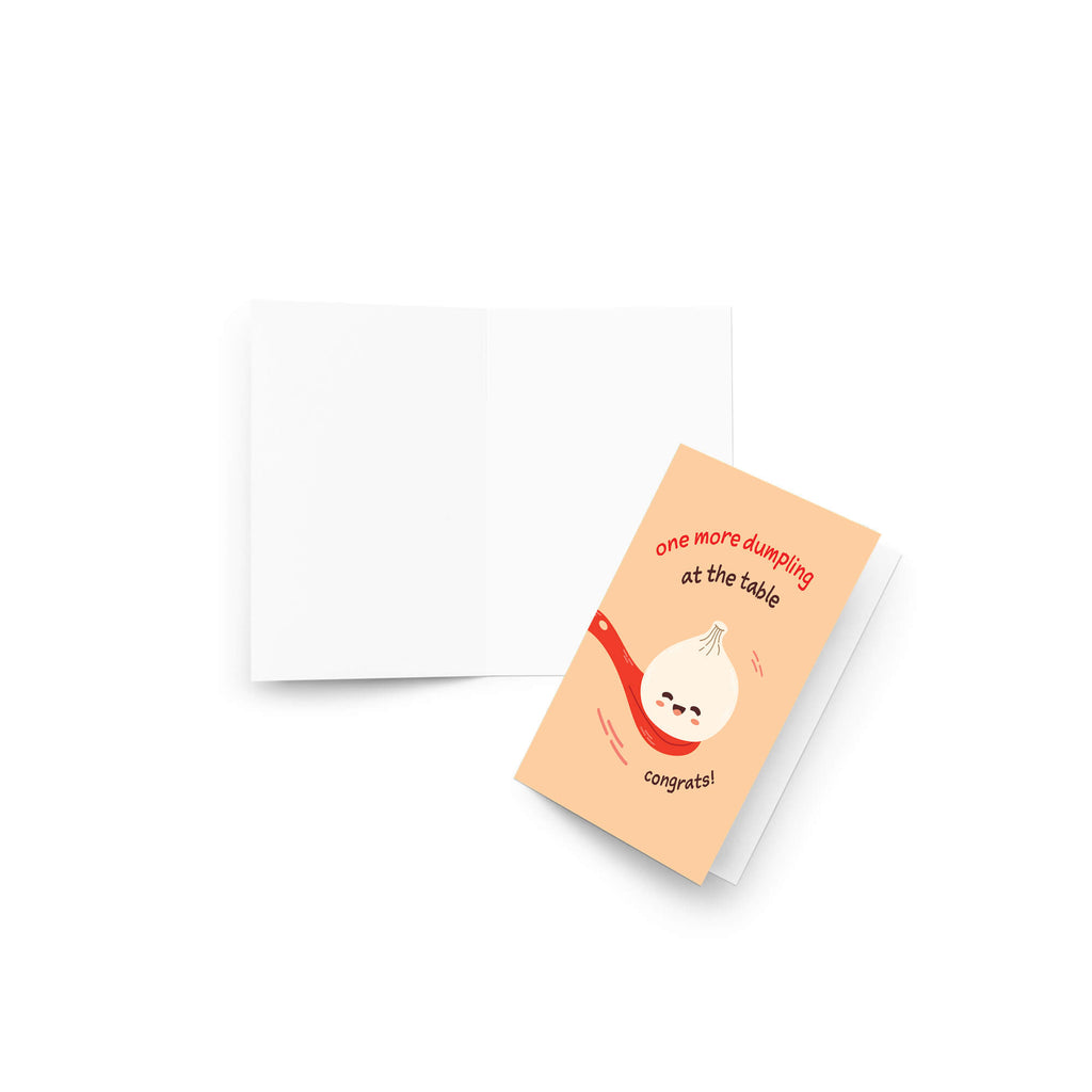 A folded newborn card with a peach front and a white back, shown on a clean white background. The front features a cartoon dumpling character with a red striped scarf and the text 'one more dumping at the table congrat!'.