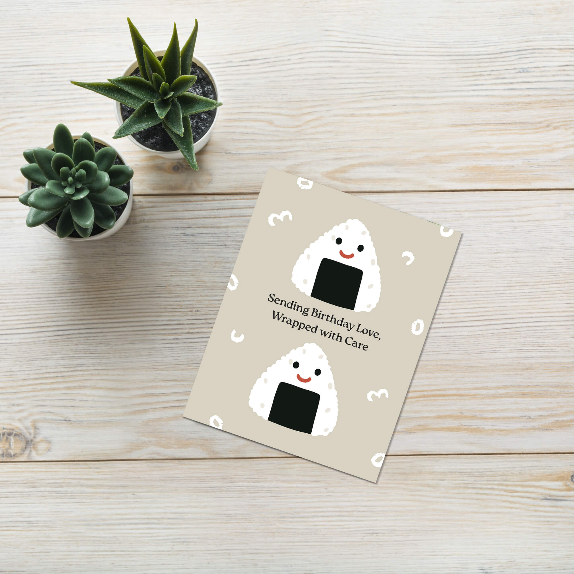 Greeting card with cartoon japanese onigiri rice ball design on a wooden surface with small potted plants.