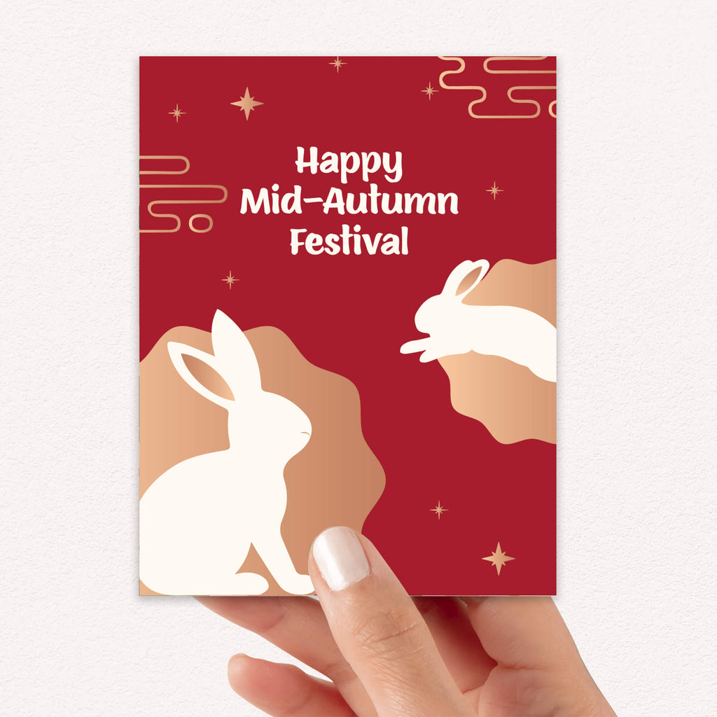 asian inspired greeting Card with white rabbits and 'Happy Mid-Autumn Festival' text on a red background, held by a hand.