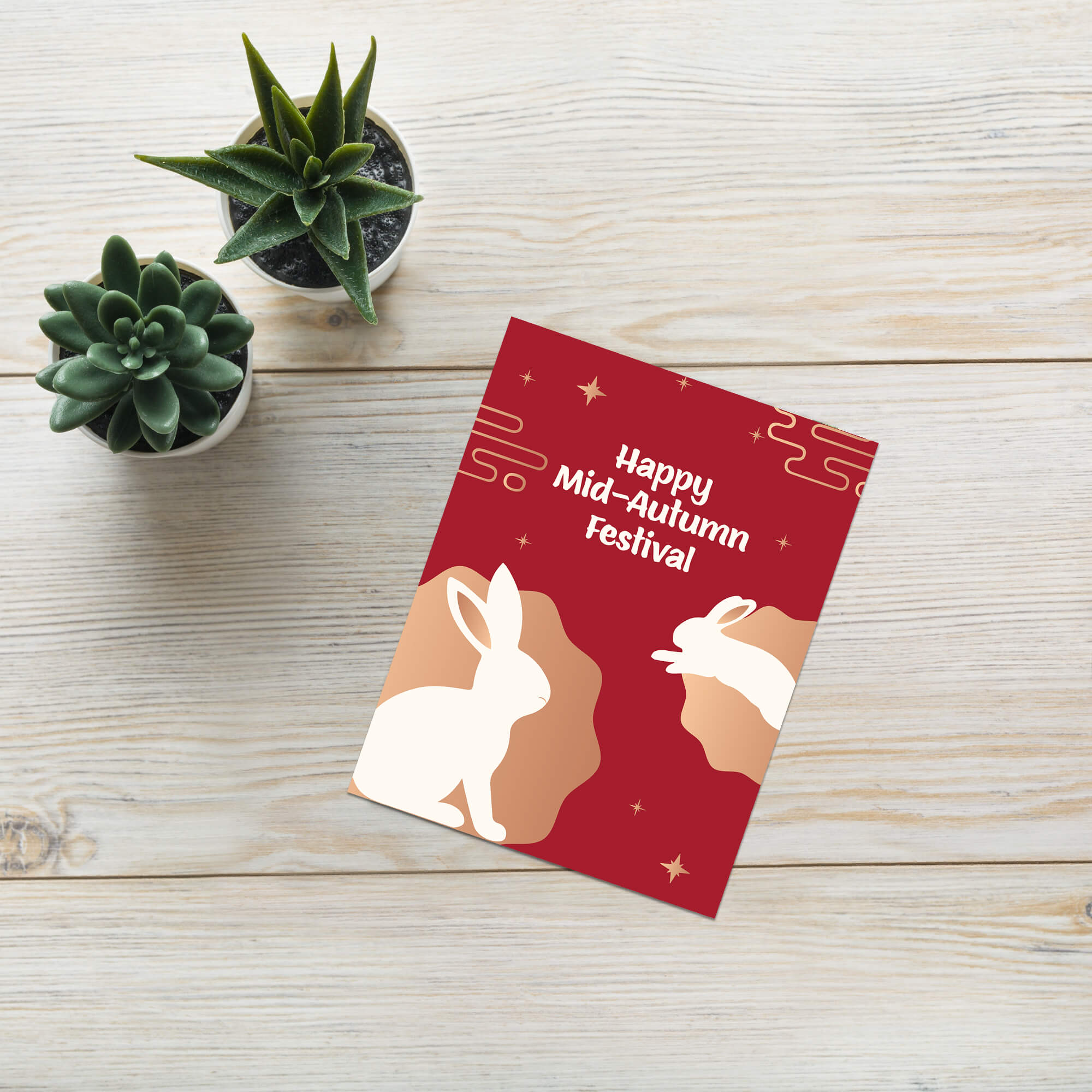 Red greeting card with white rabbits and 'Happy Mid-Autumn Festival' text on a wooden surface with small plants.