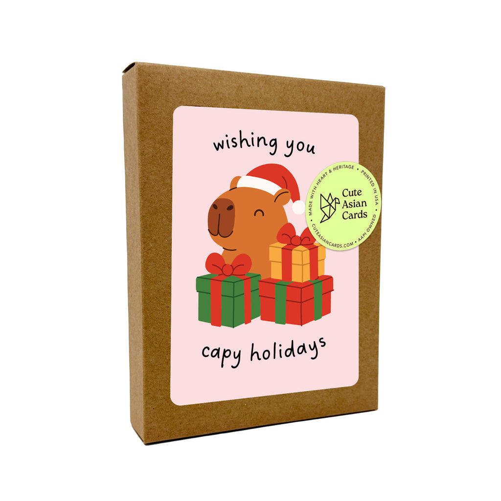 Colorful Capy Holidays card pack showing 4 holiday designs with capybaras in Santa hats, wrapped in string lights or sitting with gift boxes. Each design appears twice for a total of 8 cards with kraft envelopes.