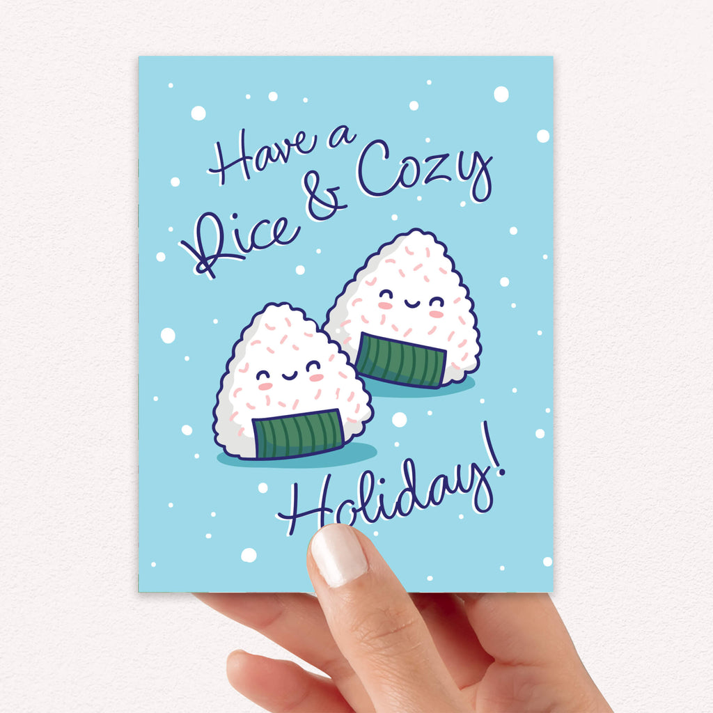unique christmas card with Two smiling onigiri (japanese rice ball snacks) on snowy blue with text “Have a Rice & Cozy Holiday.”