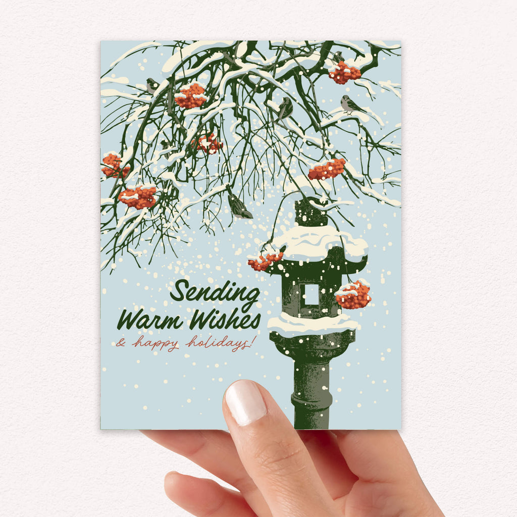Greeting card with winter scene, snow-covered branches, Snowy ishi-dōrō stone lantern with red berries, and text 'Sending Warm Wishes & happy holidays!'.