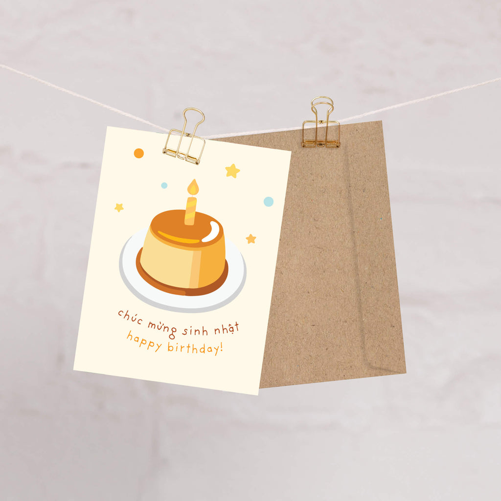 A Vietnamese birthday card clipped to a string with a kraft envelope behind it. The card features an illustration of a birthday cake with a candle and the text 'chúc mừng sinh nhật happy birthday!'.