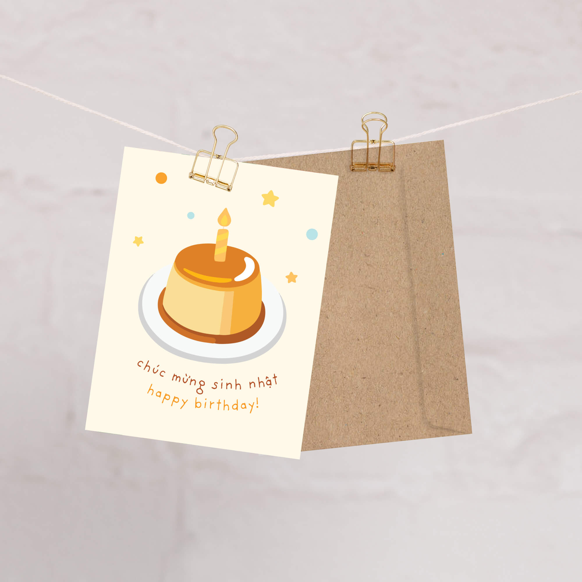 A Vietnamese birthday card clipped to a string with a kraft envelope behind it. The card features an illustration of a birthday cake with a candle and the text 'chúc mừng sinh nhật happy birthday!'.