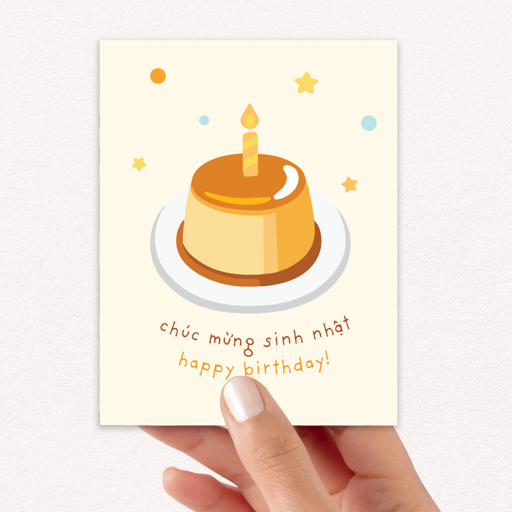 A hand holding a Vietnamese birthday card featuring a cute illustration of a vanilla custard cake with a single candle on a white plate, surrounded by small stars and dots. The card has Vietnamese and English text reading 'chúc mừng sinh nhật happy birthday!' in brown and orange lettering on a cream-colored background.