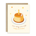 A Vietnamese birthday card with a kraft envelope, featuring an illustration of a caramel-colored flan or custard dessert on a white plate with a single candle, surrounded by small stars and dots, with the text 'Đơch mưng sinh nhát happy birthday!' in brown and orange lettering.
