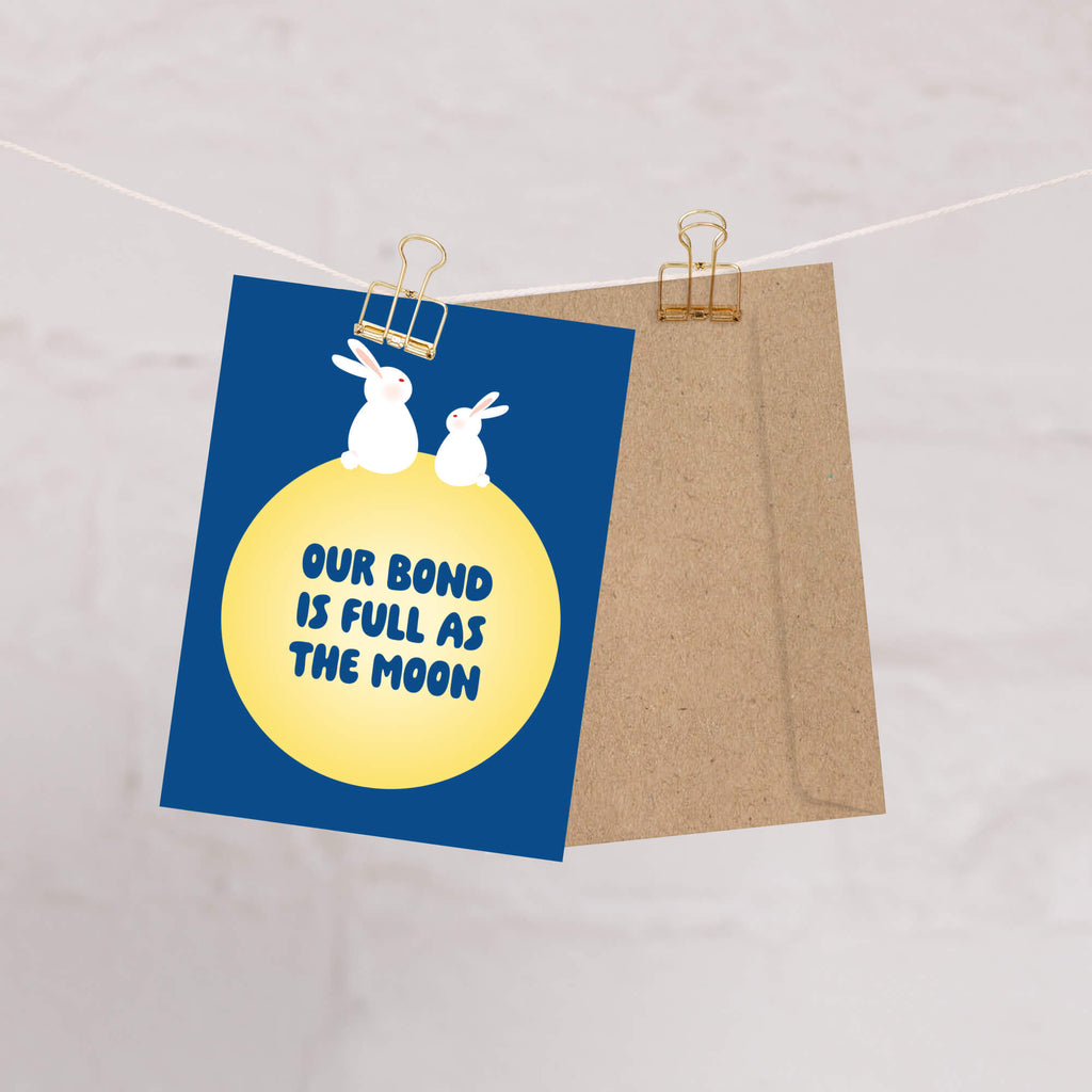 blue asian Greeting card with two rabbits on a yellow moon and 'Our bond is full as the moon' text on a blue background for mid autumn festival tsukimi or chuseok 
