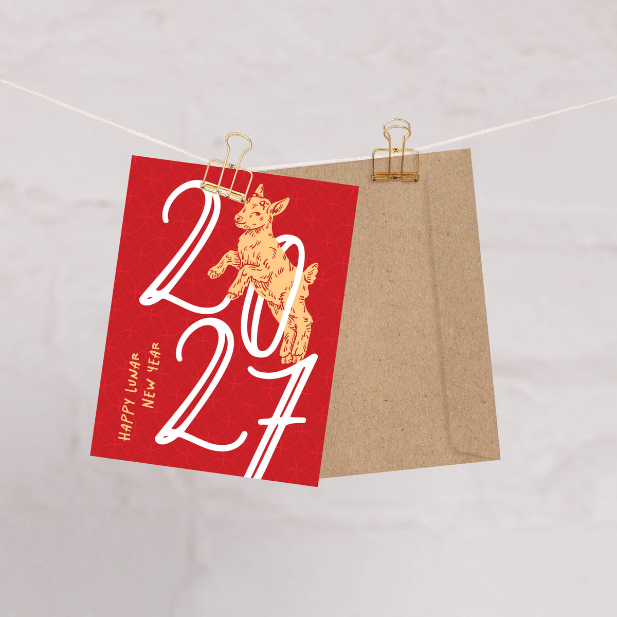 A red year of the goat 2027 card clipped to a string with a kraft paper envelope behind it, on a light neutral background. The card features a gold rabbit illustration and the text '2027', 'Happy Lunar New Year', and 'Rabbit'.