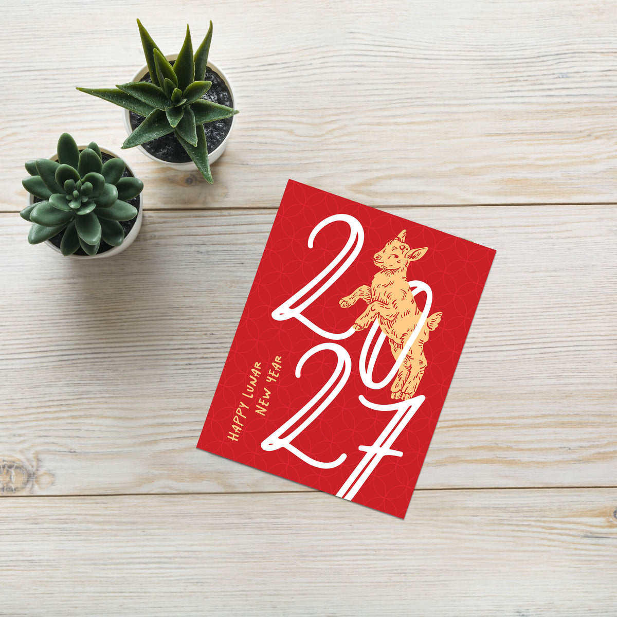 A year of the goat lunar New Year 2027 greeting card featuring a gold cat illustration and white '2027' numerals, styled on a light wood surface with two small potted succulents.