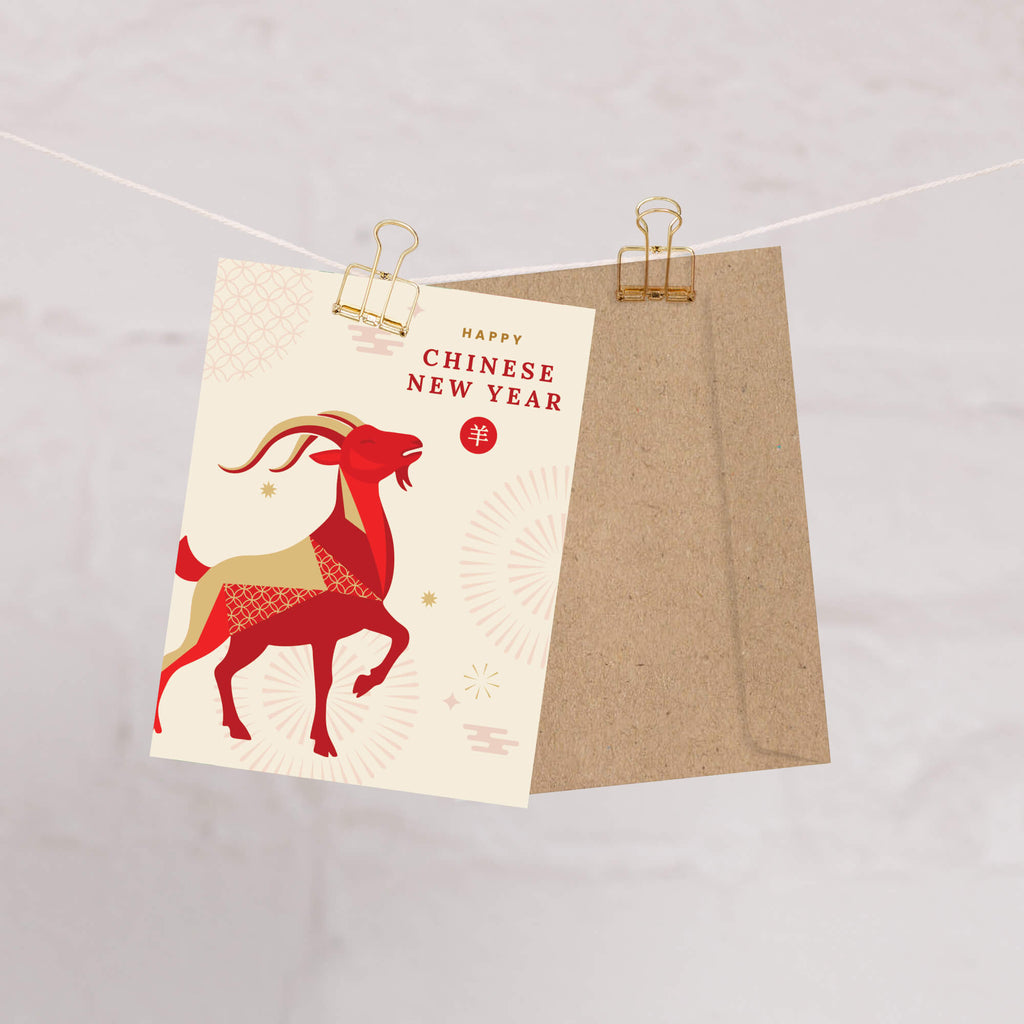 A Chinese New Year card clipped to a string with a kraft envelope behind it. The card features a red and beige stylized deer illustration with the text 'HAPPY CHINESE NEW YEAR' and a red circular emblem with a gold character.