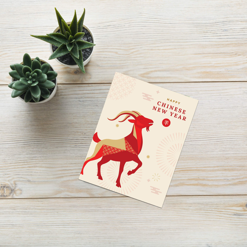 A Lunar New Year card featuring a red and gold goat illustration with the text 'Happy Chinese New Year' and a red circular seal, placed on a light wood surface next to two small potted succulents.