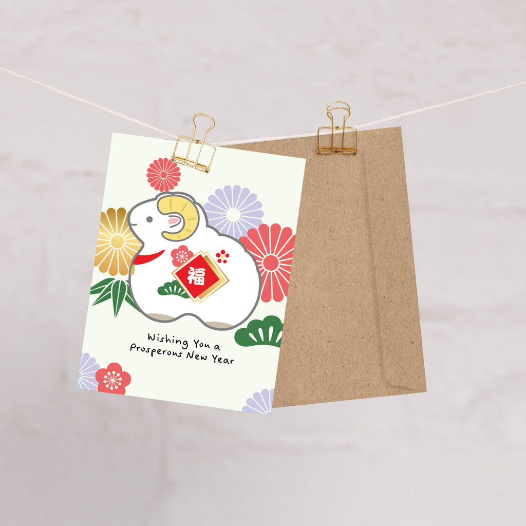 A Chinese New Year greeting card 2027 with a white ram illustration, colorful flowers, and the text 'Wishing You a prosperous New Year,' clipped beside a brown kraft envelope on a string against a light marble-like background.