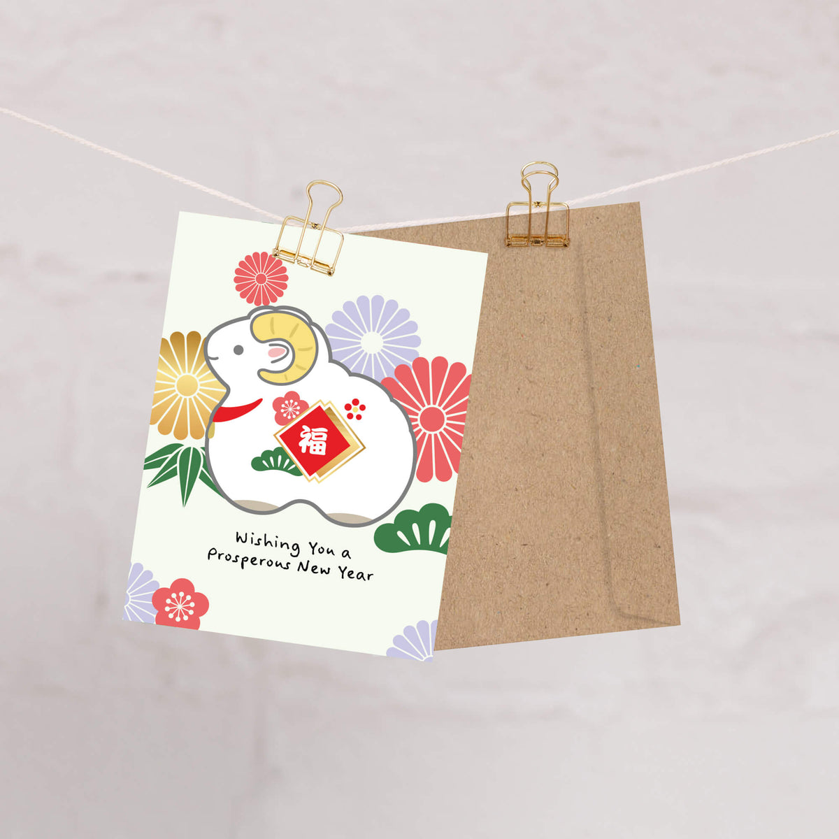 A Chinese New Year greeting card 2027 with a white ram illustration, colorful flowers, and the text 'Wishing You a prosperous New Year,' clipped beside a brown kraft envelope on a string against a light marble-like background.
