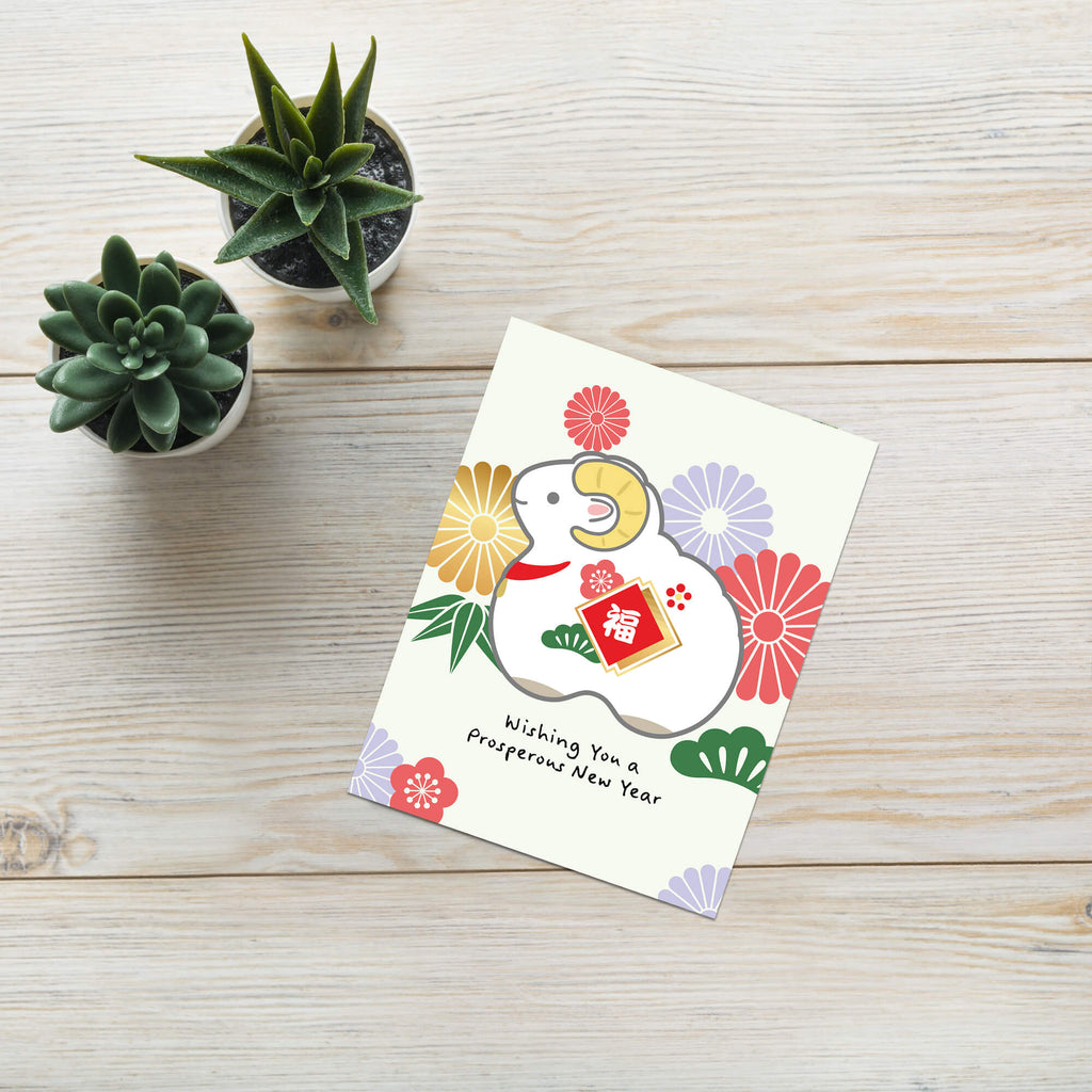 A Chinese New Year greeting card featuring a white ram surrounded by colorful flowers and a red envelope, placed on a light wood tabletop next to two small potted succulents.