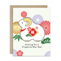 A Lunar New Year greeting card featuring a cartoon white goat with yellow horns and a red Chinese-style福 symbol on its chest, surrounded by colorful flowers and green leaves. The card reads 'Wishing You a Prosperous New Year' and is shown with a brown kraft envelope behind it on a white background.